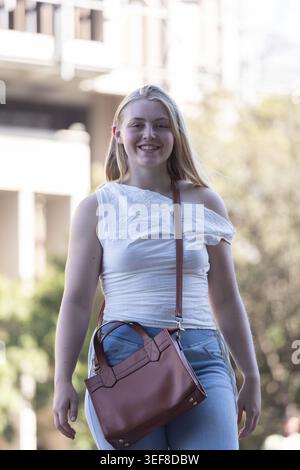 Woman walking toward camera on campus wearing white ruched top, light-blue jeans, brown leather bag Stock Photo