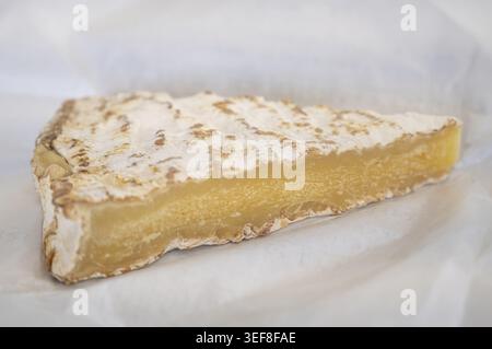 A cut piece of Brie de Meaux aged for six months with a distinct straw-yellow tint of the dough and velvety white mold bark on light wrapping paper in Stock Photo