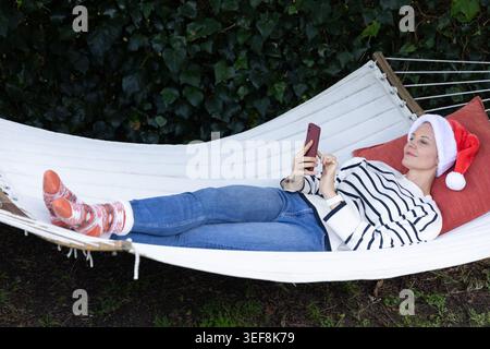 Woman reclining on white hammock in garden wearing Santa hat and striped top, using smartphone Stock Photo