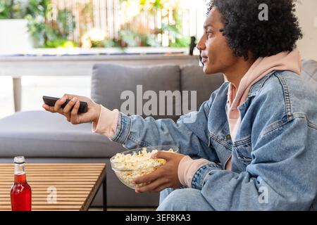 African American man sitting on gray sofa at home holding TV remote and popcorn, copy space Stock Photo