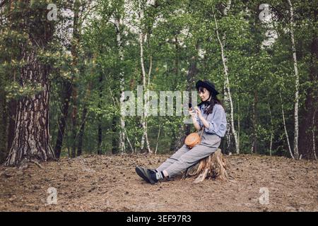 Person wearing hat sits on tree stump in forest and looks at smartphone. AI chatbot for everyday use, virtual assistant in nature, smart AI lifestyle. Stock Photo