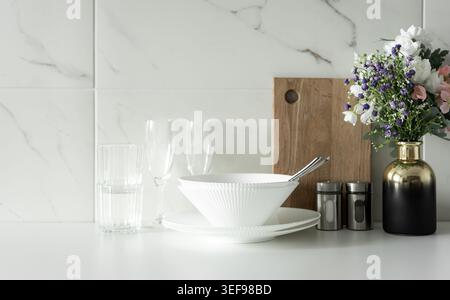 Minimal white table setting with bowls, glasses and flowers on kitchen counter in bright modern interior. Stock Photo