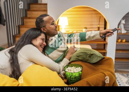 Happy smiling multiracial couple cuddled on a cozy sofa, sharing popcorn and a remote while watching a movie or show in their relaxed, intimate living Stock Photo