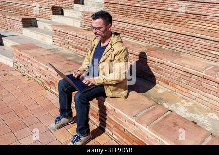 Man typing on a laptop computer while sitting outside on brick amphitheater steps, representing flexible remote work and digital nomadism Stock Photo