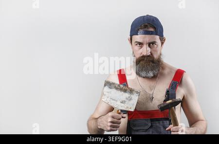 Bearded man in overalls and a baseball cap holds a hammer and spatula. Portrait of a worker. Copy space for your text Stock Photo
