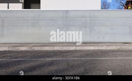 Gray plaster striped fence with white house on behind. Concrete sidewalk and street in front. Background for copy space. Stock Photo
