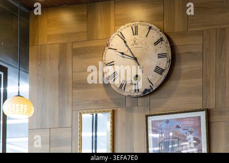A large antique round clock with Roman numerals and weathered face hangs prominently on a natural wood-paneled wall, flanked by a warm pendant lamp and framed art, creating a nostalgic rustic cafe atmosphere. Stock Photo