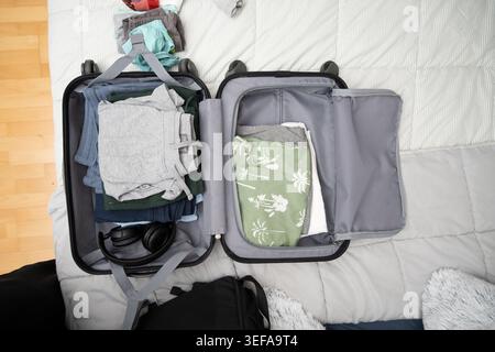Open suitcase with clothes, accessories, and headphones on a bed preparing for a trip Stock Photo