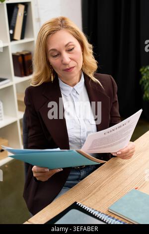 Mature woman in a chic blazer examines important papers with confidence at her workspace. Stock Photo