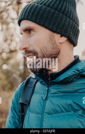 Sideways shot of unshaven man with dark thick beard, wears warm har and anorak, looks pensively aside, poses outside, dreams about something has outdoor stroll in unknown place alone, active lifestyle Stock Photo