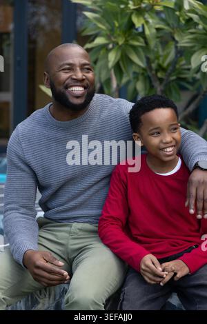 African American father and child sitting on stone ledge at patio, wearing grey sweater, wristwatch. Family, outdoor, greenery, embrace, bond, warmth, casual Stock Photo