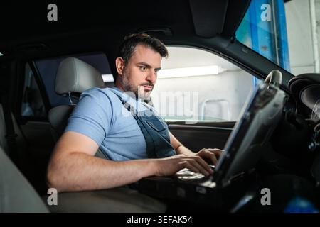 Auto mechanic using laptop computer while sitting in car and working on vehicle diagnostics in a repair shop Stock Photo