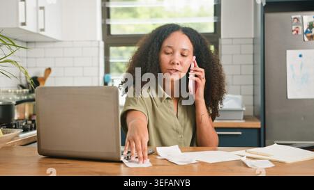 Woman Working From Home Kitchen Paying Bills On Phone While Using Laptop And Documents Stock Photo