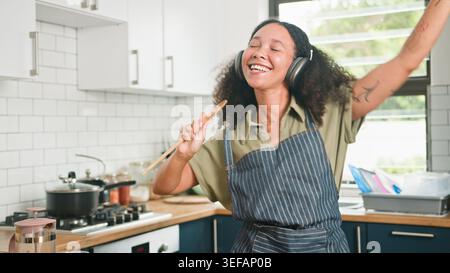 Joyful Woman Cooking In Kitchen Wearing Headphones And Apron Singing While Preparing Meal Stock Photo