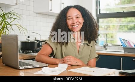 Smiling Woman Working on Laptop in Bright Kitchen With Receipts and Notebook Stock Photo