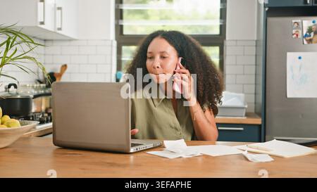 Young Woman Working From Home At Kitchen Table On Laptop While Talking On Phone And Reviewing Bills Stock Photo
