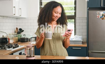 Smiling Woman Relaxing With Coffee And Smartphone In Modern Kitchen While Making French Press Stock Photo