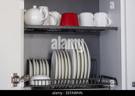 A kitchen dish rack stores an assortment of white ceramic plates and bowls on the bottom shelf, while the top shelf holds several white mugs, a teapot Stock Photo
