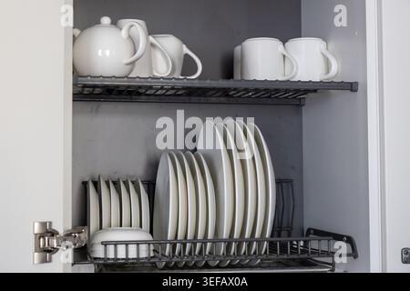 A kitchen dish rack stores an assortment of white ceramic plates and bowls on the bottom shelf, while the top shelf holds several white mugs, a teapot Stock Photo