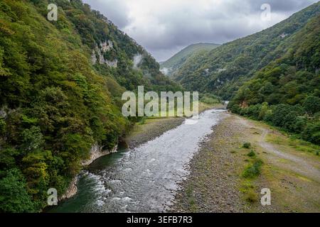 Lupshara river flows through a green valley in Ritsa Relict National Park with rocky banks and forested hills in Russia Stock Photo