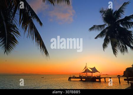 Beautiful sunset over the ocean near Coco de Mer hotel in Seychelles with silhouettes of palm trees and a wooden pier Stock Photo