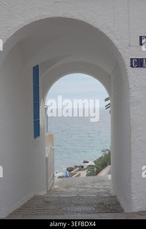 Mediterranean Sea viewed through a whitewashed arched passageway with mosaic tile steps and a blue shutter in Nerja, Andalusia, Spain. Stock Photo
