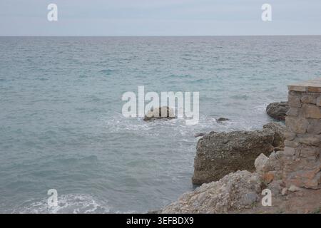 Rocky limestone coastline with ancient stone wall and gentle waves on the turquoise Mediterranean Sea under an overcast sky in Nerja, Andalusia, Spain Stock Photo