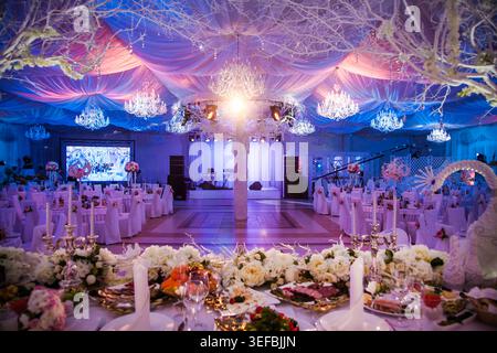Elegant event venue decorated for wedding reception. Tables set for banquet with flowers. Hall features draped ceiling, crystal chandeliers, and blue Stock Photo