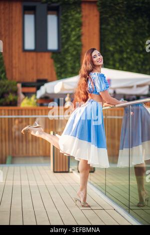 Joyful woman with long auburn hair in blue and white dress poses on modern terrace. She smiles, kicking one leg up playfully. Sunny day lighting creat Stock Photo