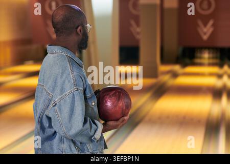 Black young adult man holding bowling ball and aiming toward lane in bowling alley, side view. Representing leisure activity and recreation marketing, medium shot Stock Photo