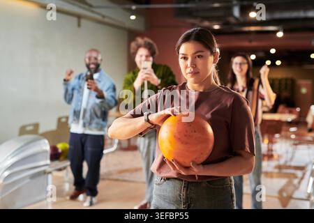 Portrait of young woman holding bowling ball while coworkers cheering in bowling alley, representing team building and office bonding. For HR culture, workplace engagement campaigns Stock Photo