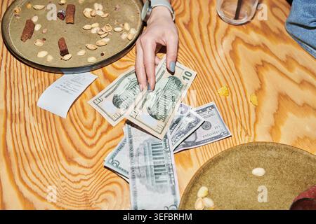 Close up of young adult woman hand placing cash on restaurant table beside receipt and plates, showing bill payment and spending. Useful for dining cost, budgeting, tipping concepts Stock Photo