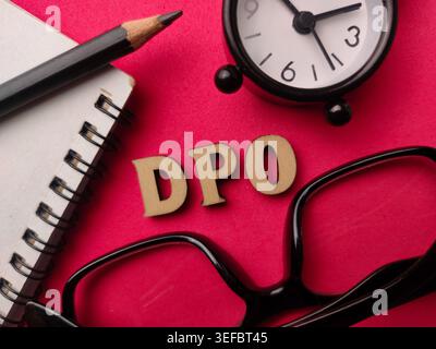 Wooden letters spelling "DPO" on a red surface with a clock, notebook, pencil, and black glasses. Stock Photo
