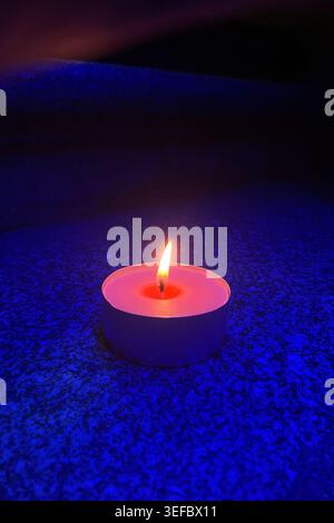 Burning tea light candle in blue night lighting. single candle flame glows orange against a dark blue grainy surface. The small flame Stock Photo