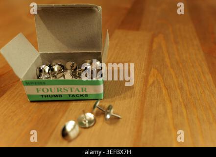 Silver metal fourstar thumb tacks spilling from an open cardboard box, resting on a clean wooden desk surface, presenting office and stationery suppli Stock Photo
