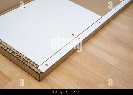 White wooden furniture panels sit inside an open cardboard shipping box on a laminate floor. Stock Photo