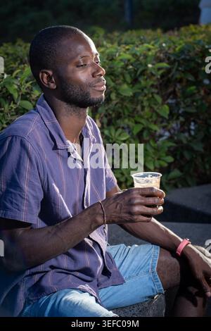 Relaxed young Black man drinking iced coffee outdoors in sunlight during casual urban lifestyle moment with authentic candid expression. Stock Photo