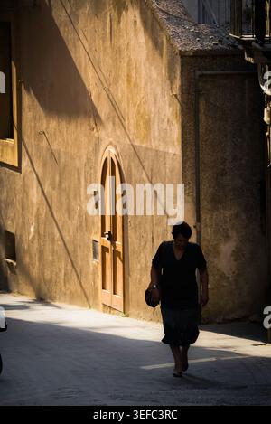 Lonely woman, view of a middle aged female walking alone in shadow in an empty street in the historic old town centre of Enna, Sicily. Stock Photo