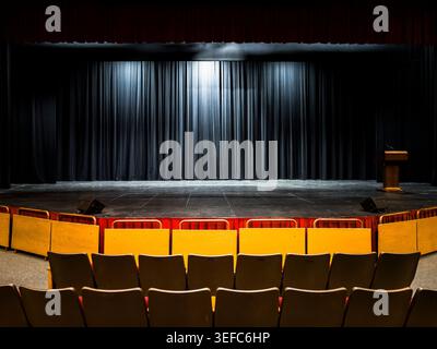 An empty dark theater stage with a wooden podium wooden partitions and rows of unoccupied seats under a spot lit black velvet curtain background Stock Photo