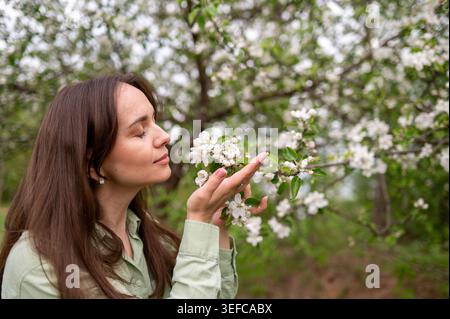 A side view portrait of a happy young caucasian woman smelling delicate white blossoms on an apple tree branch while relaxing in a beautiful blooming Stock Photo