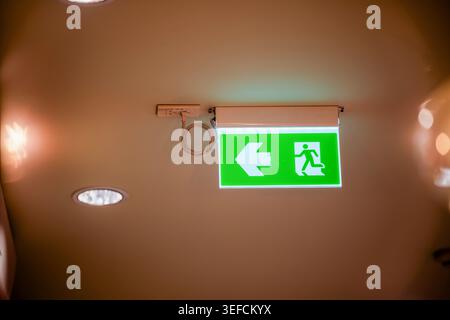 A bright green emergency exit sign mounted on a ceiling features a running figure and arrow, clearly indicating the emergency exit route in public or Stock Photo