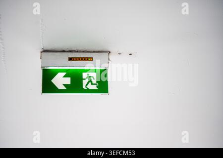 A green illuminated emergency exit sign with a left arrow and running figure, indicating the direction for evacuation in case of emergencies, mounted Stock Photo