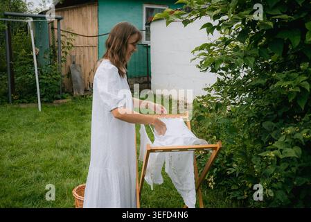 Young smiling brunette woman in white dress hanging wet clean white laundry on wooden clothes drying rack in backyard garden near village house Stock Photo