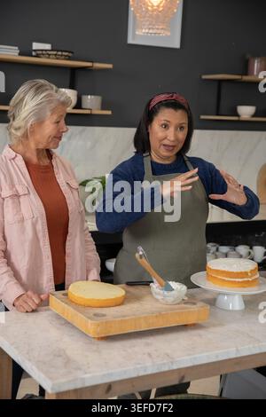 Diverse senior women in kitchen wearing gray apron, pink shirt, preparing layered cake with spatula. Baking, dessert, decorating, frosting, lesson, co Stock Photo
