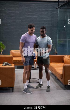 Sharing workout stats, two men in activewear checking smartphone in gym lounge with blue bottle. Fitness, partners, conversation, hydration, technolog Stock Photo