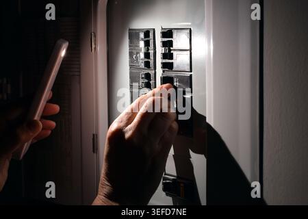 Closeup of person using smartphone flashlight while switching electrical circuit breakers in dark home. Residential electricity, power outage, emergen Stock Photo