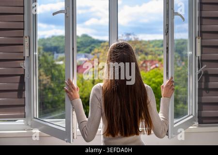 Girl looking outside through open window. Teen girl seen from behind opening a large window and enjoying sunlight, fresh air, and green outdoor Stock Photo