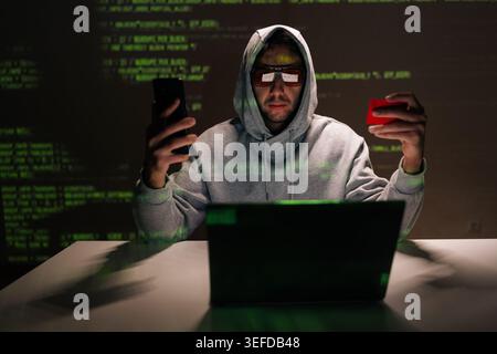 Portrait of hooded hacker in glasses sitting at desk, staring at smartphone while holding red credit card, laptop open and green code reflecting on Stock Photo