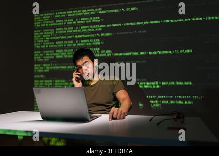 Programmer sitting at desk with laptop, deeply engaged in phone conversation while analyzing projected green code, symbolizing complex software Stock Photo