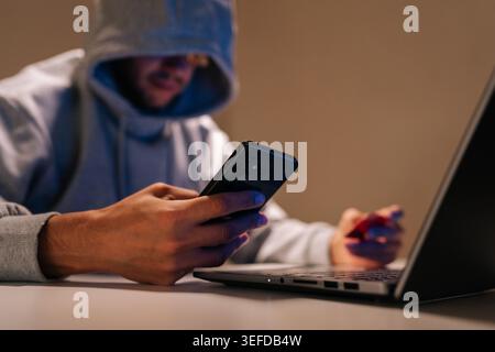 Close-up of hooded person engaging in cybercrime, performing online fraud and data theft using mobile phone, laptop and credit card in dark Stock Photo